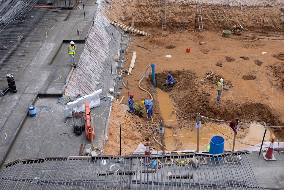 Cove Edition I By Imtiaz 1 Construction workers are digging in a site with exposed soil, machinery, and tools for foundation work and water management.