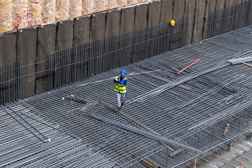 Cove Edition I By Imtiaz 1 A construction worker in a blue hard hat and reflective vest stands on a steel rebar structure at a site.