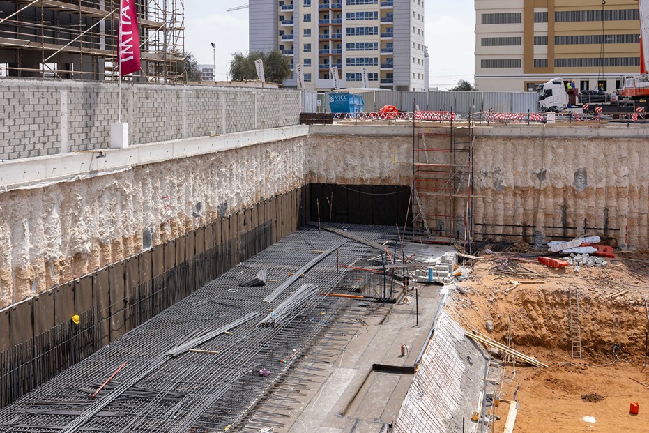 Cove Edition I By Imtiaz 1 Construction site with exposed foundation, steel rebar, scaffolding, and nearby buildings under development.