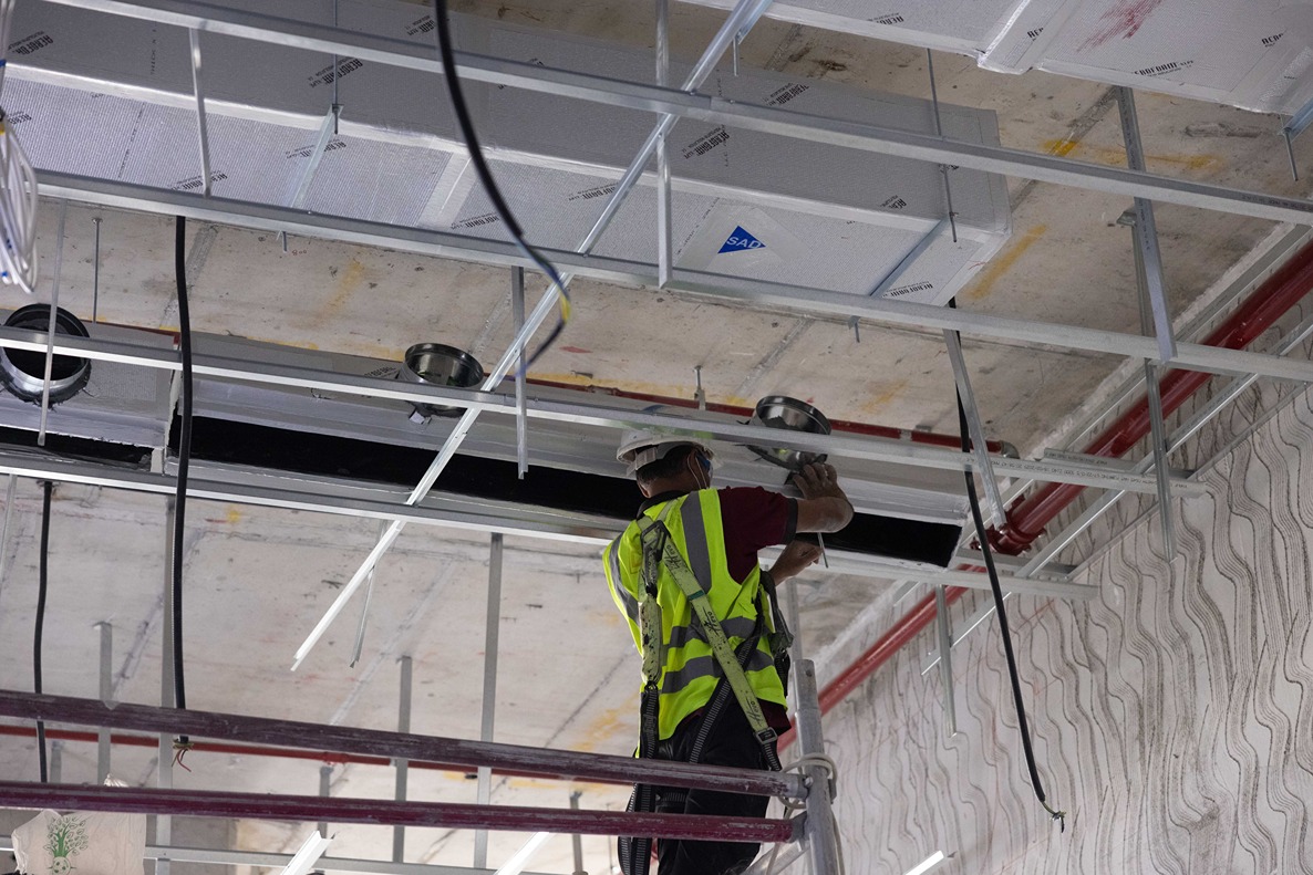 Beach Walk Residence A worker in a safety vest installs fixtures on a ceiling while standing on scaffolding in a construction site.