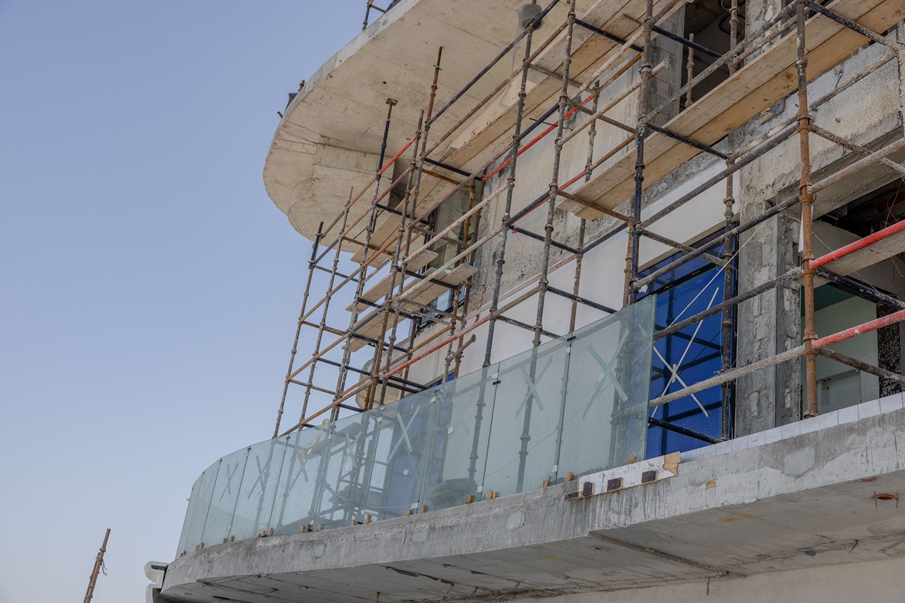 Beach Walk Residence Construction site showing scaffolding on a building with a glass balcony railing and unfinished walls against a blue sky.