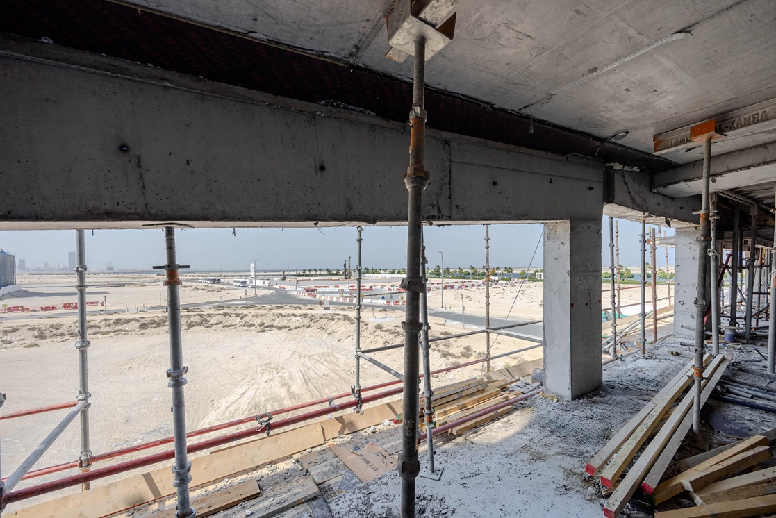 Beach Walk Iii By Imtiaz Construction site view through scaffolding, showing sandy terrain and distant buildings under a clear sky.