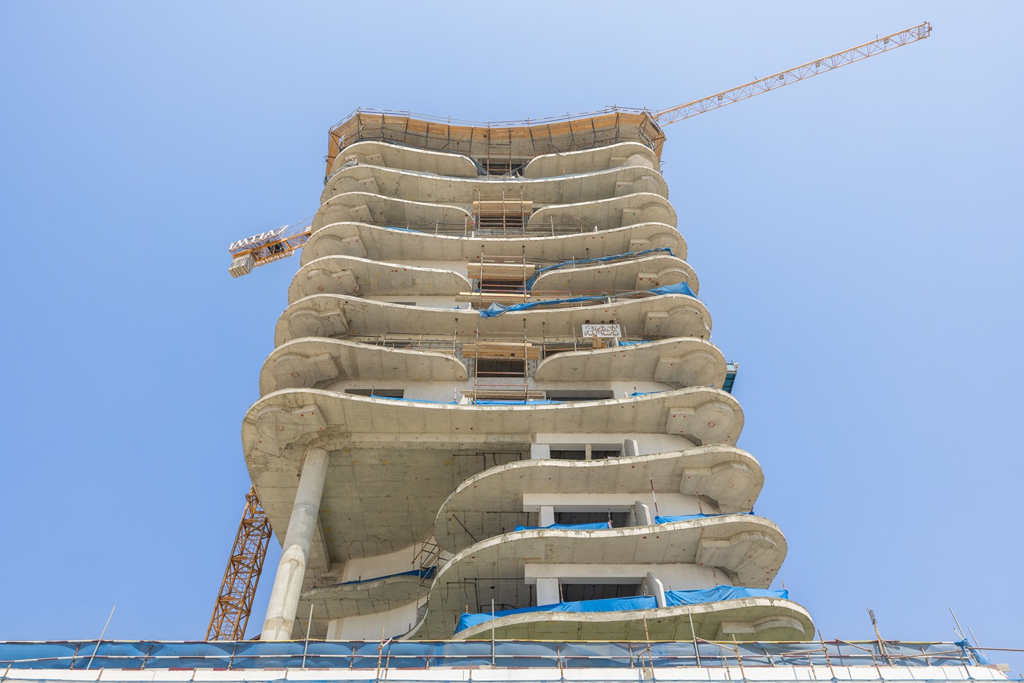 Beach Walk Residence A tall building under construction with a unique wavy design and scaffolding, set against a clear blue sky.