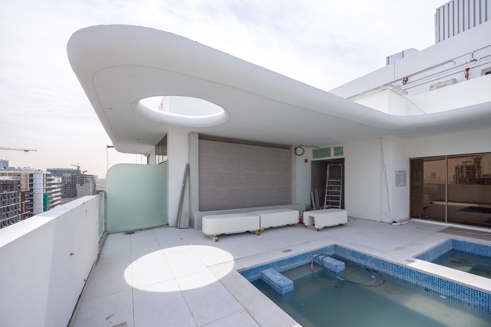 Pearl House By Imtiaz A modern balcony with white seating, a pool, and a sleek canopy overlooking a cityscape under a cloudy sky.