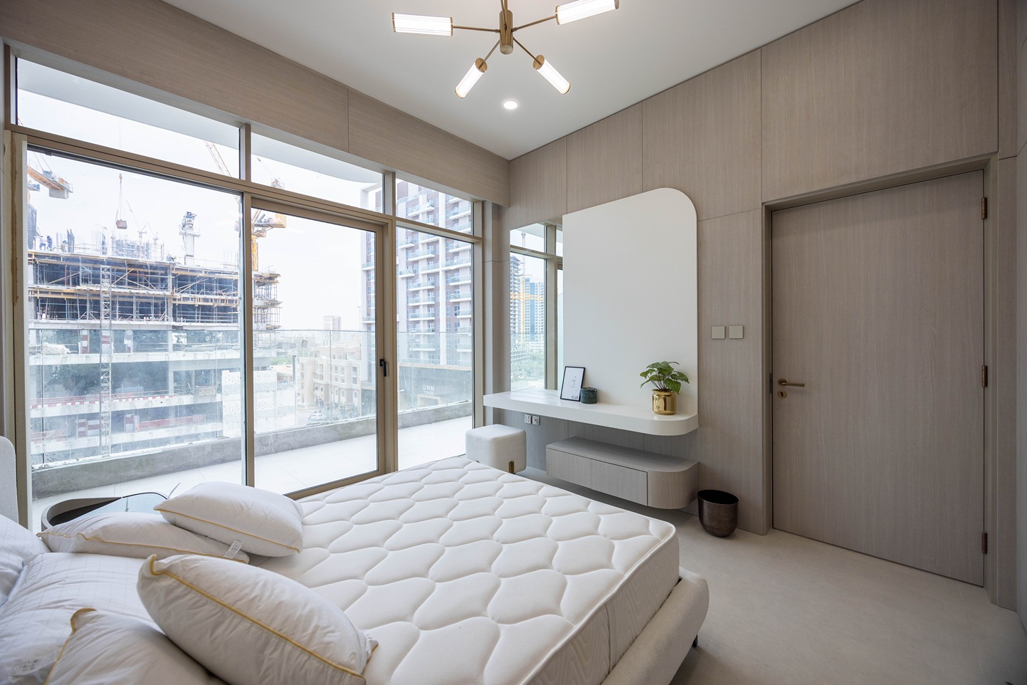 Pearl House By Imtiaz A modern bedroom featuring a bed with white bedding, a desk, and large windows overlooking a construction site.