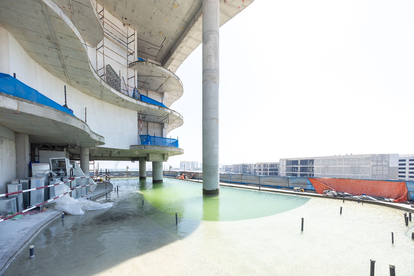 Beach Walk Residence An under-construction building with curved concrete architecture, surrounded by a green water pond and scaffolding.