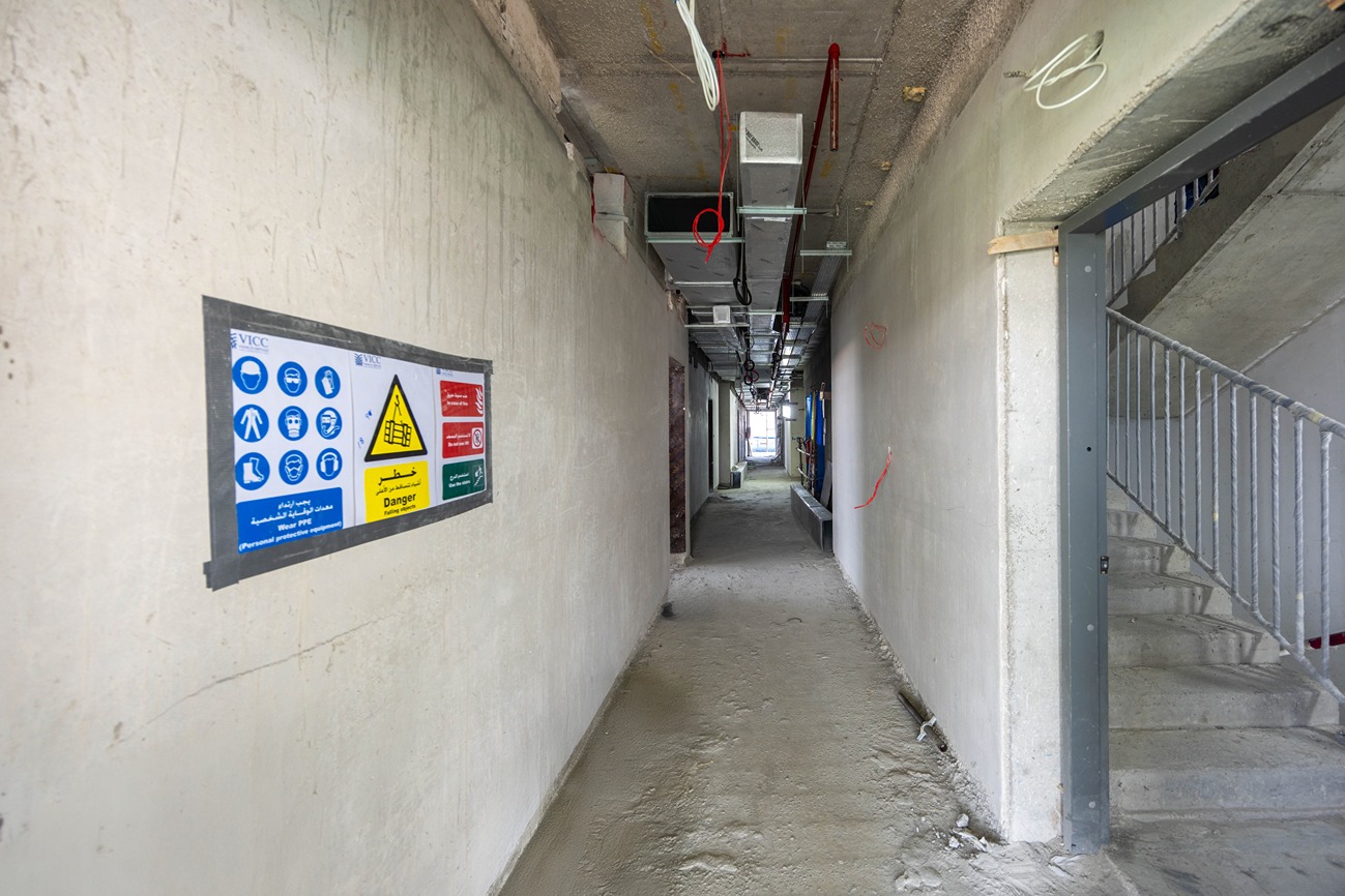 Beach Walk Residence A narrow construction corridor with walls, pipes above, and safety signs posted on the left side.