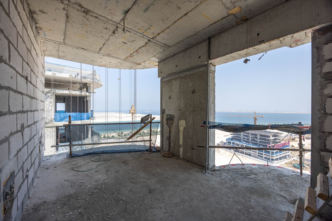 Beach Walk Residence Under-construction interior space with a view of cranes and buildings by the sea, showcasing a stark concrete environment.