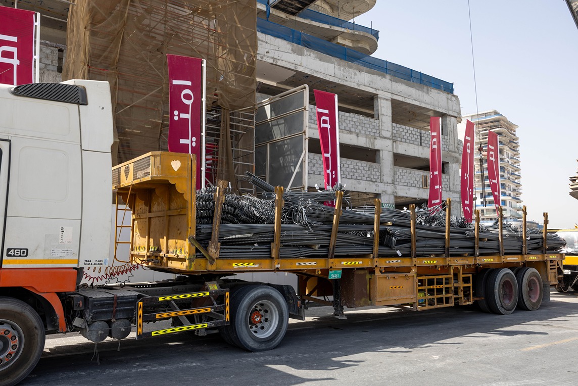 Beach Walk Iii By Imtiaz A truck loaded with steel bars parked next to a construction site, featuring banners and scaffolding.