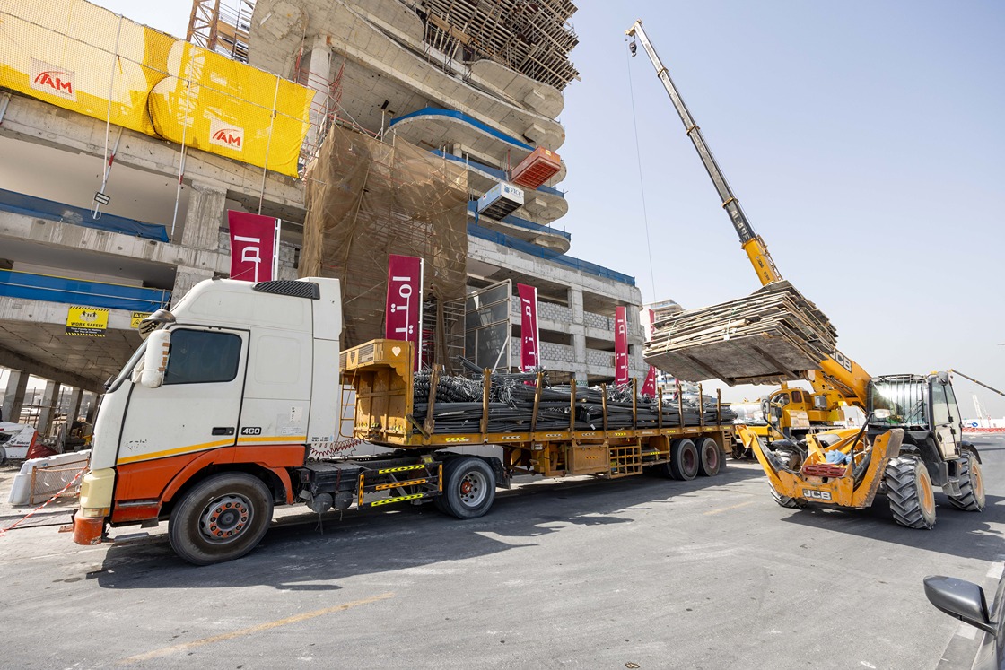 Beach Walk Iii By Imtiaz A construction site with a truck unloading materials and a crane lifting wooden planks near a building structure.