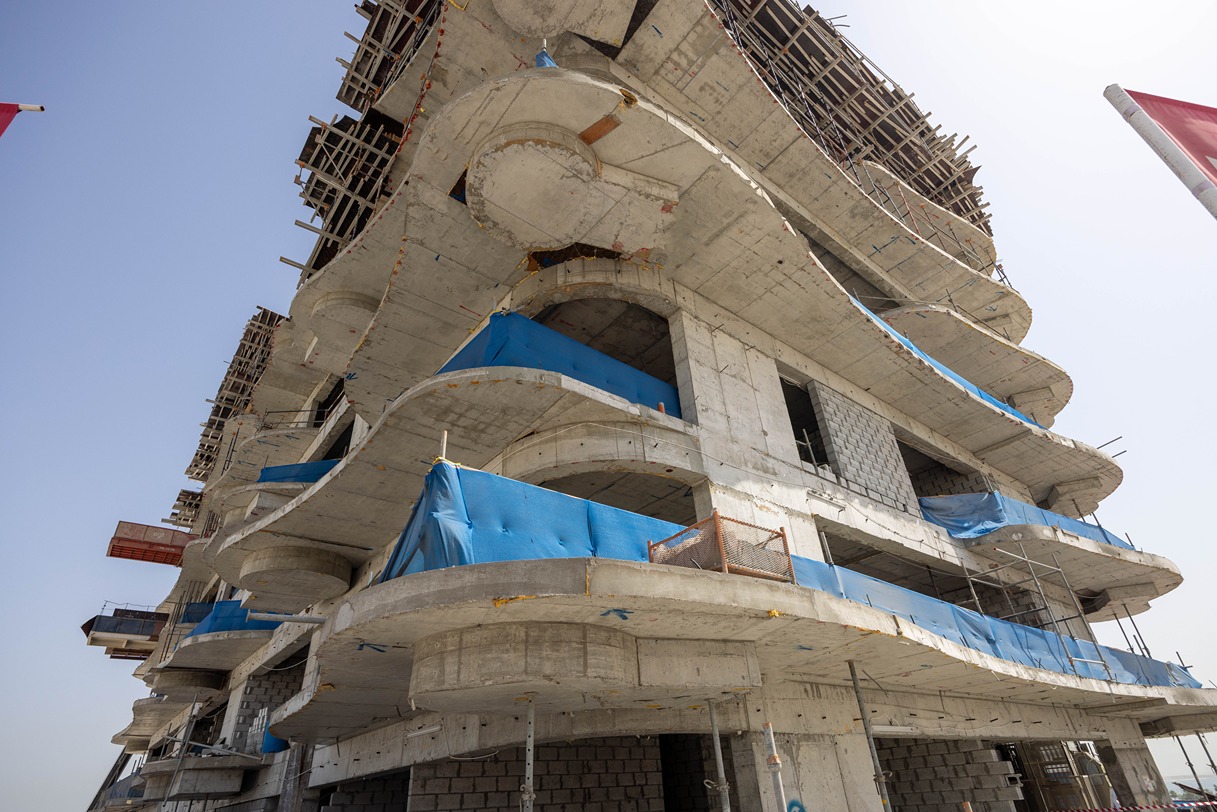 Beach Walk Iii By Imtiaz A partially constructed building features curved concrete elements and blue construction tarps against a clear sky.