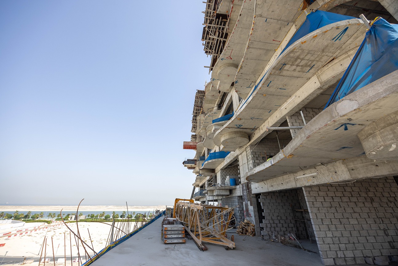 Beach Walk Iii By Imtiaz Construction site with concrete structures, scaffolding, and equipment, overlooking a coastline and blue sky.