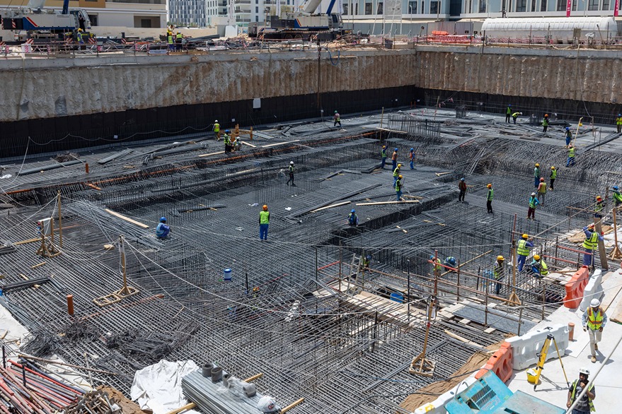 Cove Edition I By Imtiaz 1 Construction site showing workers in hard hats and safety gear managing rebar and foundation work in a deep pit.