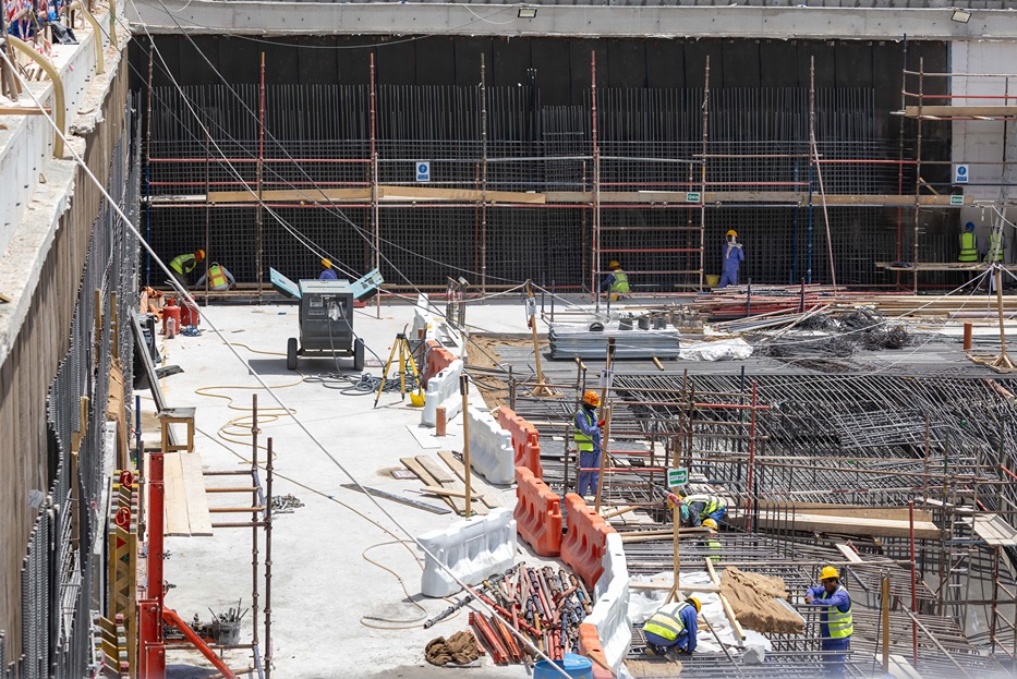 Cove Edition I By Imtiaz 1 Construction site with workers in helmets, moving materials and equipment among steel reinforcements and scaffolding.