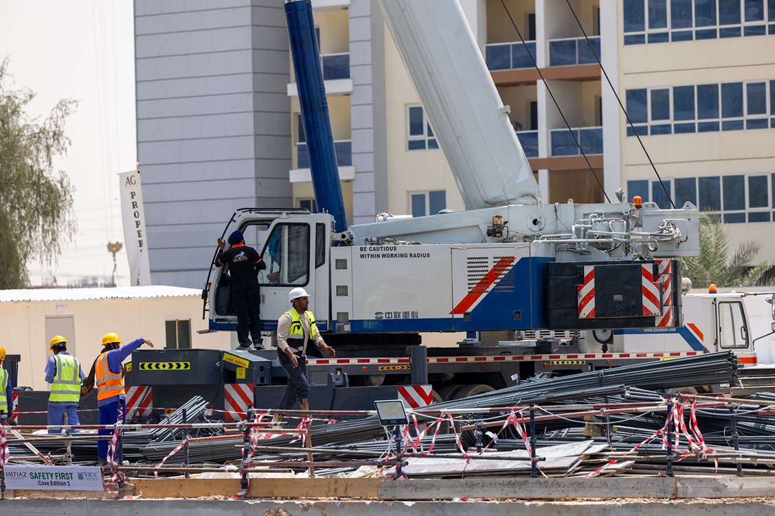 Cove Edition I By Imtiaz 1 Construction site featuring a crane, workers in safety gear, and bundles of steel rebar. Safety signs visible.