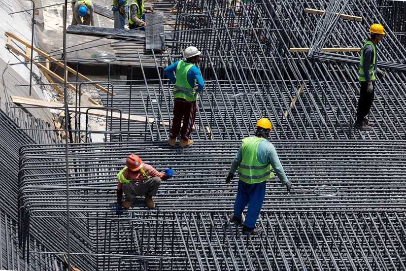 Cove Edition I By Imtiaz 1 Construction workers wearing helmets and vests manage steel rebar on a building site, with materials scattered around.