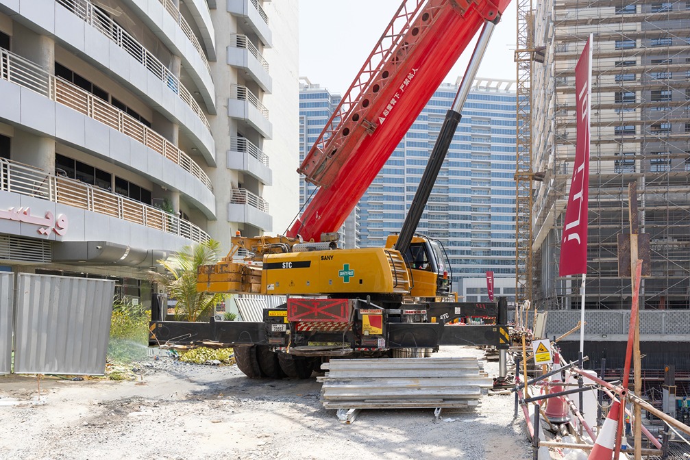 Cove Edition I By Imtiaz 1 A construction site featuring a yellow crane with a red arm, surrounded by buildings and scaffolding.