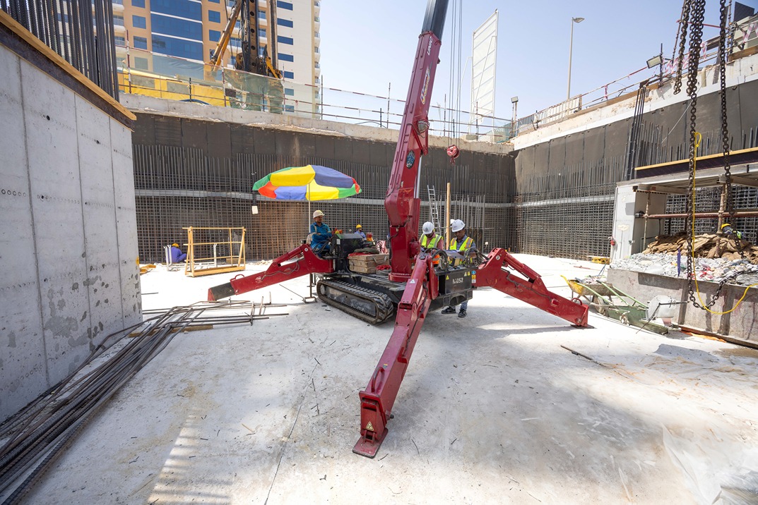 Cove Edition I By Imtiaz 1 Construction site with workers operating a red crawler crane under a colorful umbrella, surrounded by building materials.