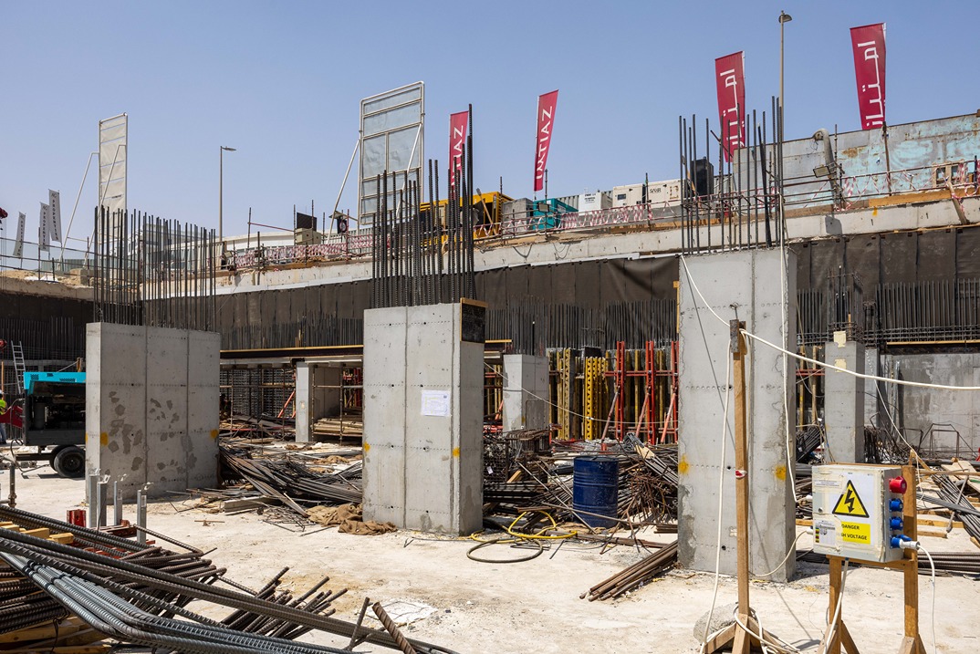 Cove Edition I By Imtiaz 1 Construction site with concrete pillars, rebar, and machinery. Flags visible atop a partially completed structure.