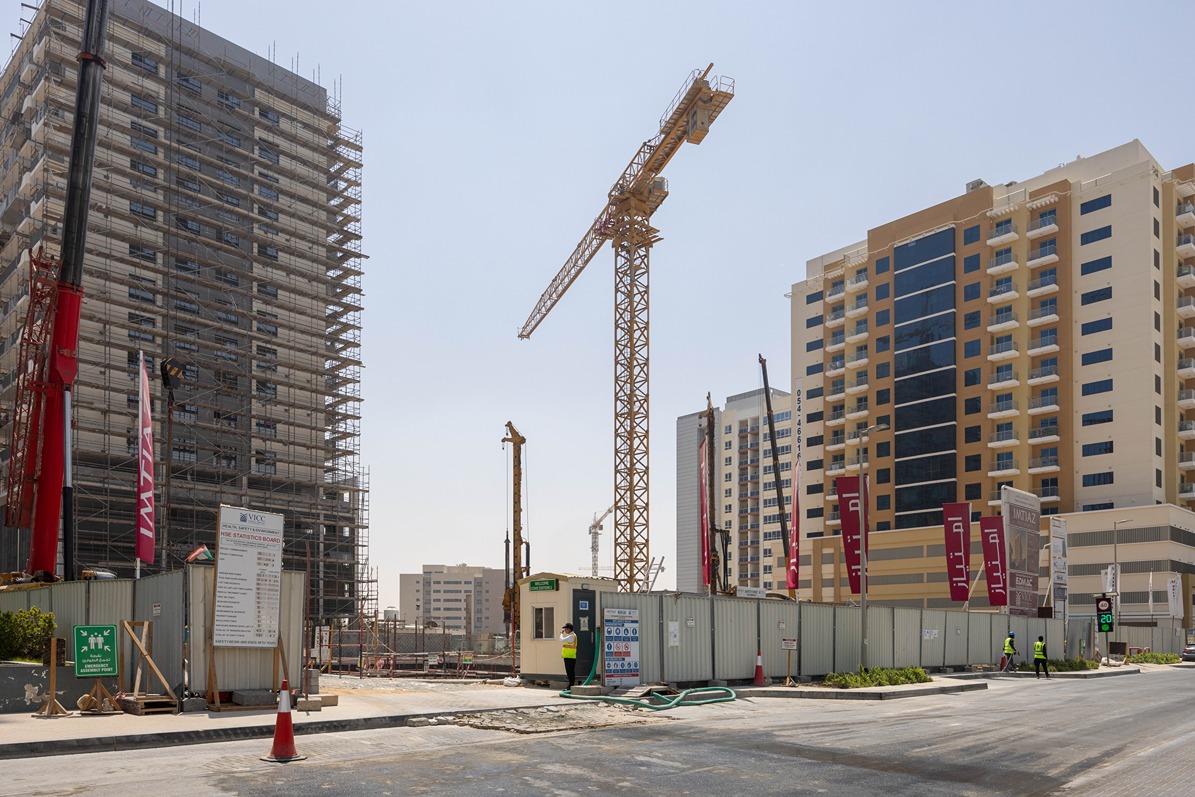 Cove Edition I By Imtiaz 1 Construction site featuring cranes, scaffolding, and workers, with nearby buildings and safety signage visible.