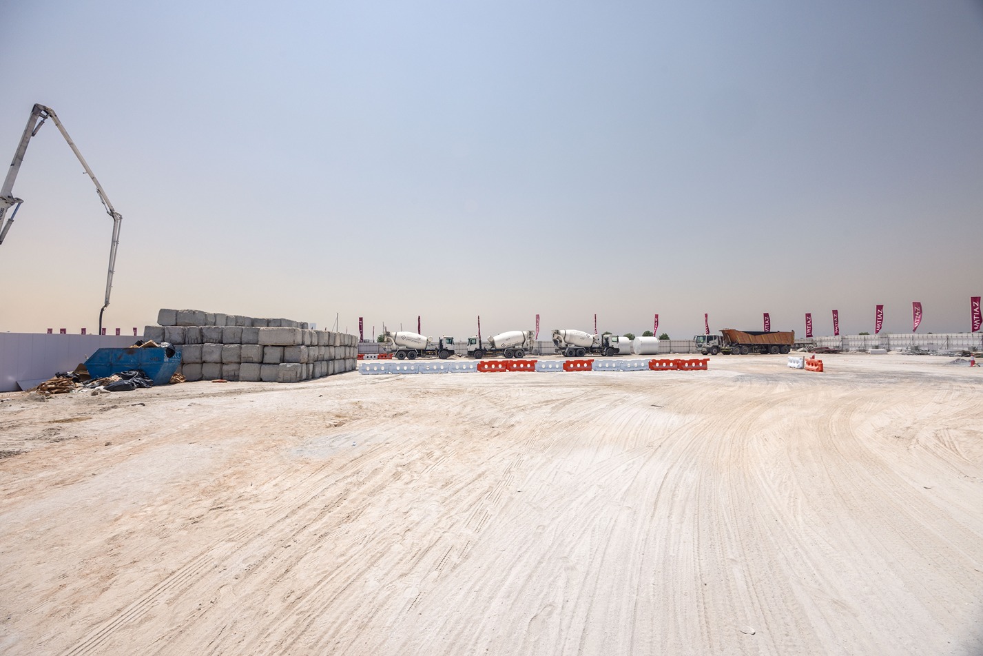 Cotier House 2 By Imtiaz Construction site with concrete trucks, stacked materials, and safety barriers on a sandy surface under a clear sky.