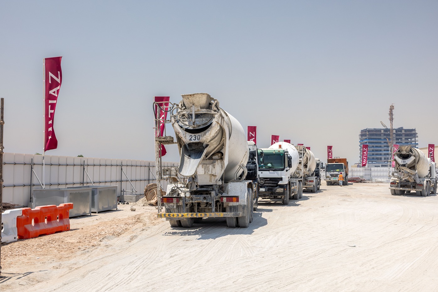 Cotier House 2 By Imtiaz A row of concrete mixer trucks on a construction site, with banners and a building in the background.
