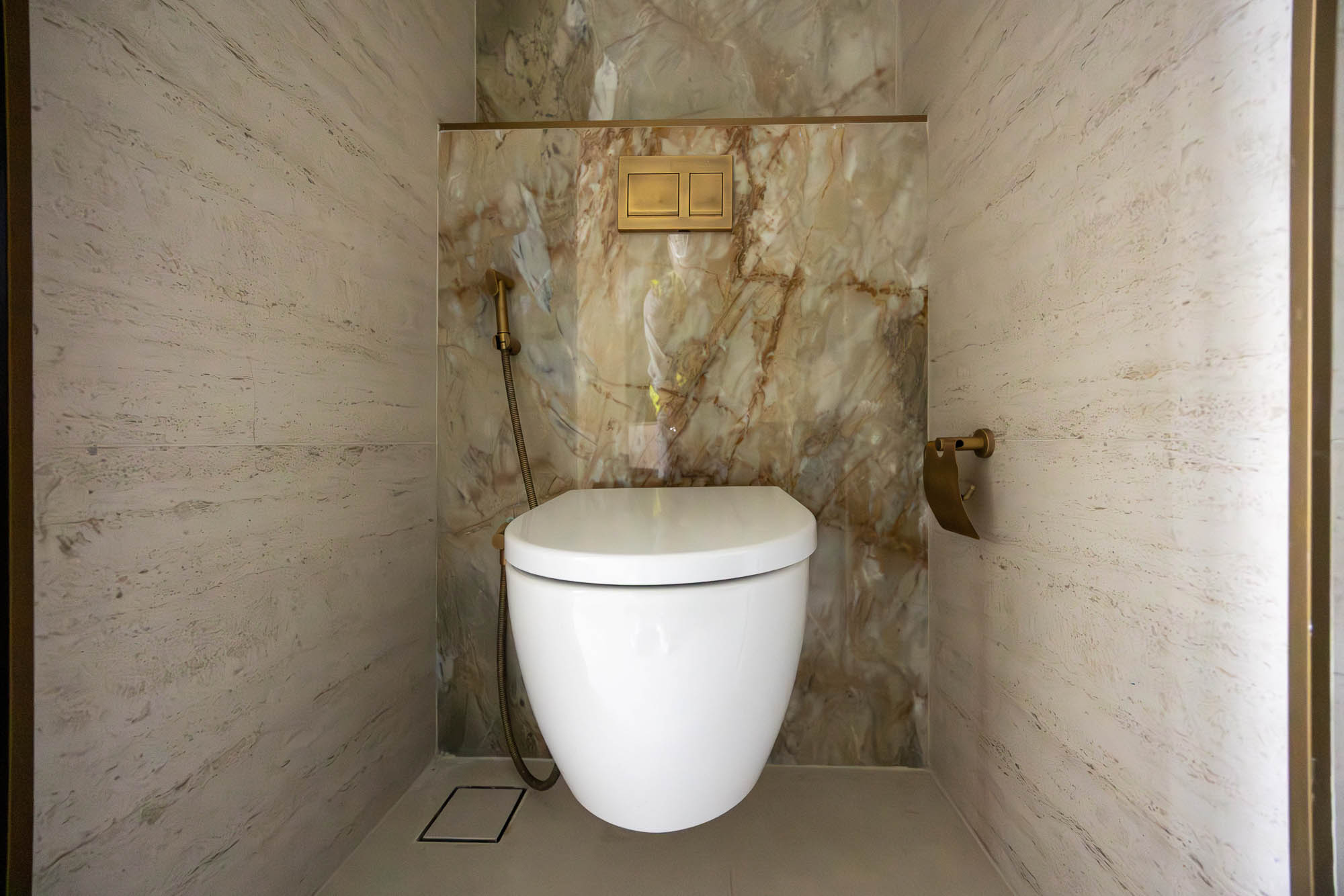 Pearl House By Imtiaz Modern bathroom featuring a white toilet, marble walls, and a gold toilet paper holder. Elegant and minimalist design.