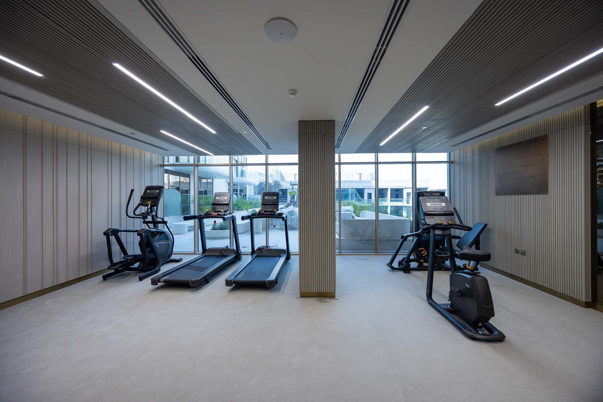 Pearl House By Imtiaz A modern gym interior features two treadmills and an elliptical machine with large windows for natural light.