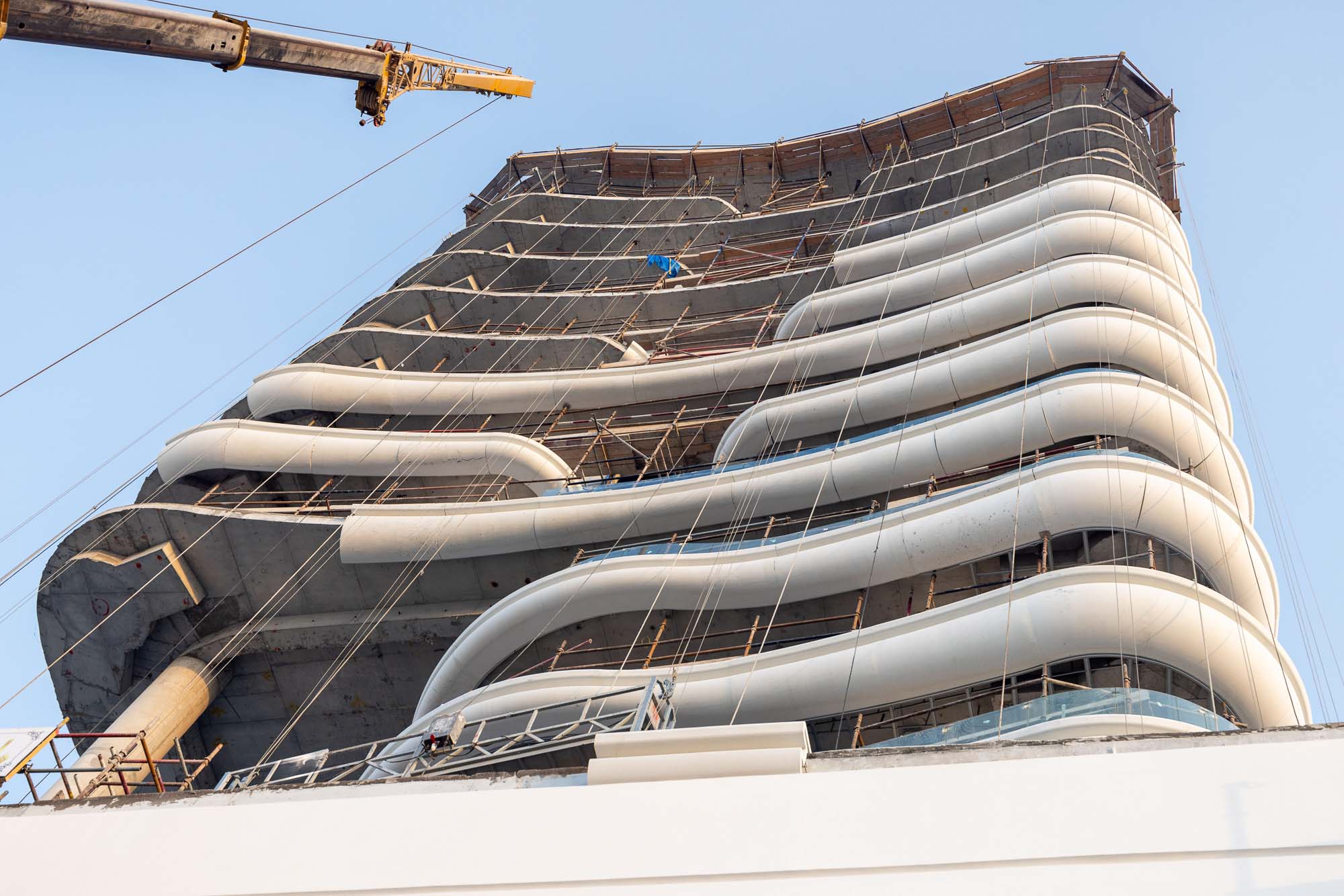 Beach Walk Residence A construction site featuring a modern building with curved balconies and a crane overhead against a clear sky.