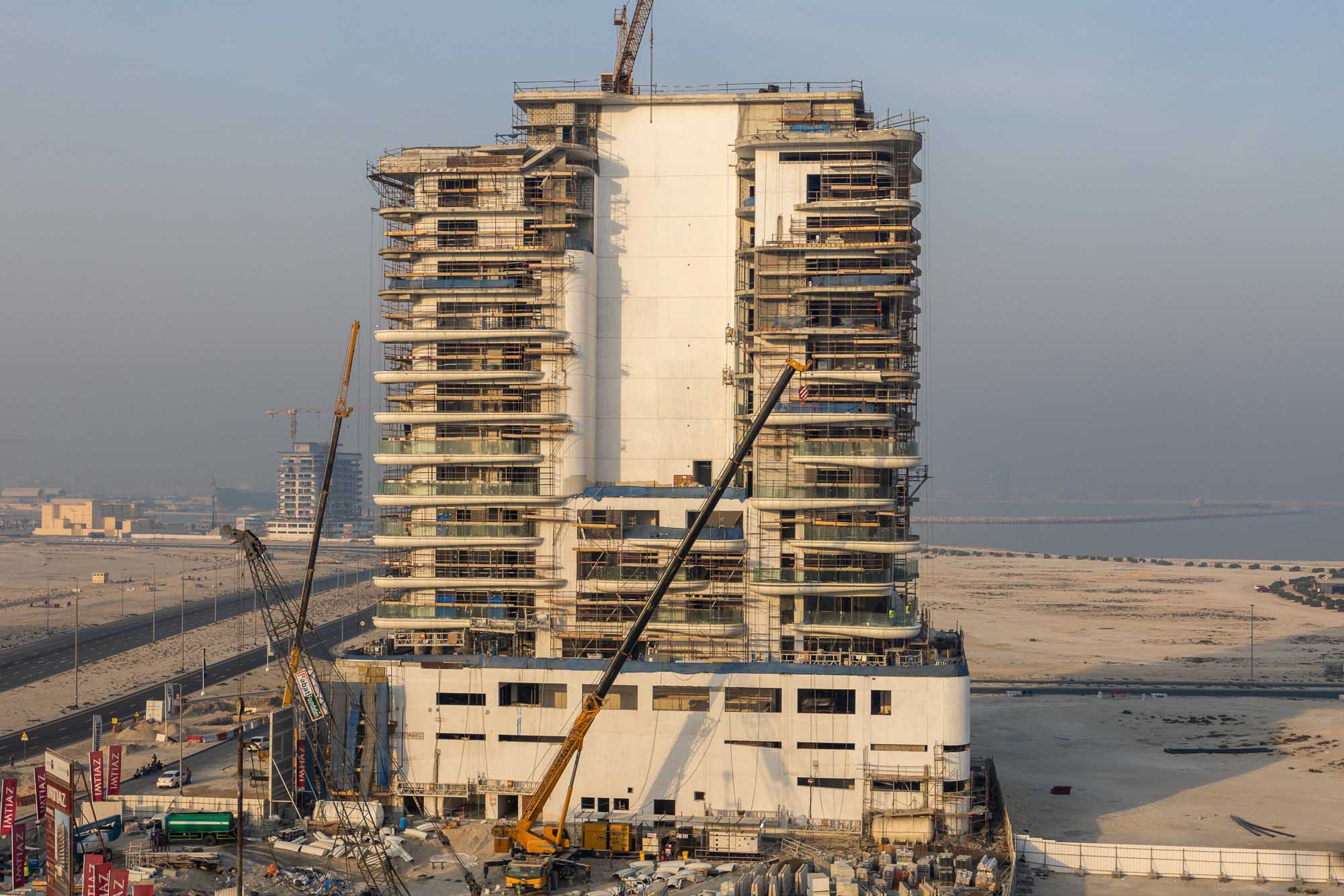 Beach Walk Residence A modern skyscraper under construction, featuring cranes and scaffolding, with a sandy landscape in the background.