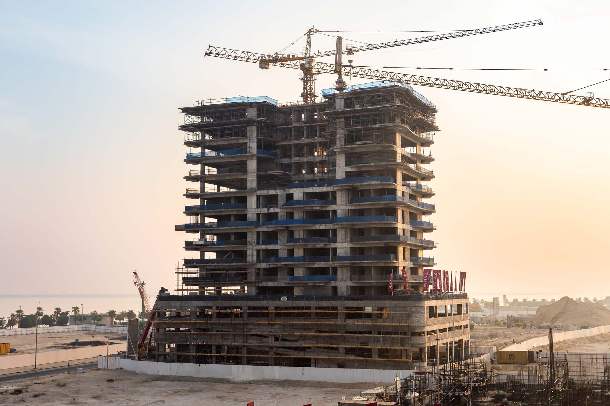 Beach Walk Iii By Imtiaz Construction site of a tall building with scaffolding, cranes, and a sandy area in the foreground at sunset.