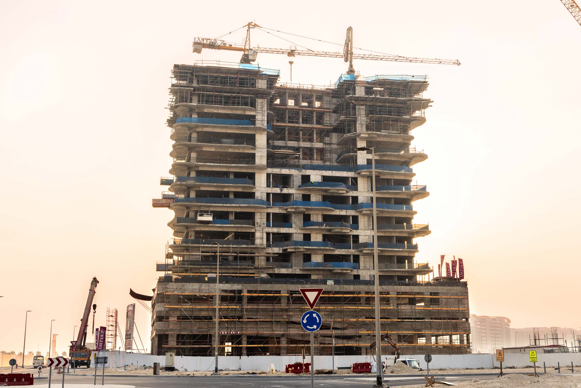 Beach Walk Iii By Imtiaz A tall building under construction with scaffolding, cranes, and nearby road signs in an urban setting.