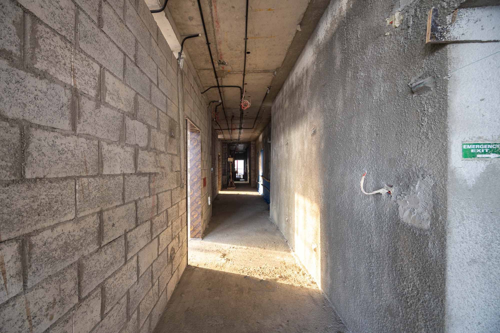 Beach Walk Iii By Imtiaz A long, unfinished hallway with concrete block walls, exposed wiring, and light streaming in from the end.