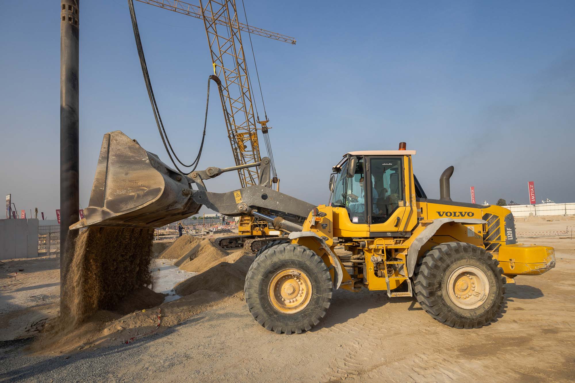 Cotier House 2 By Imtiaz A yellow Volvo wheel loader at a construction site, scooping and pouring sand with a crane in the background.