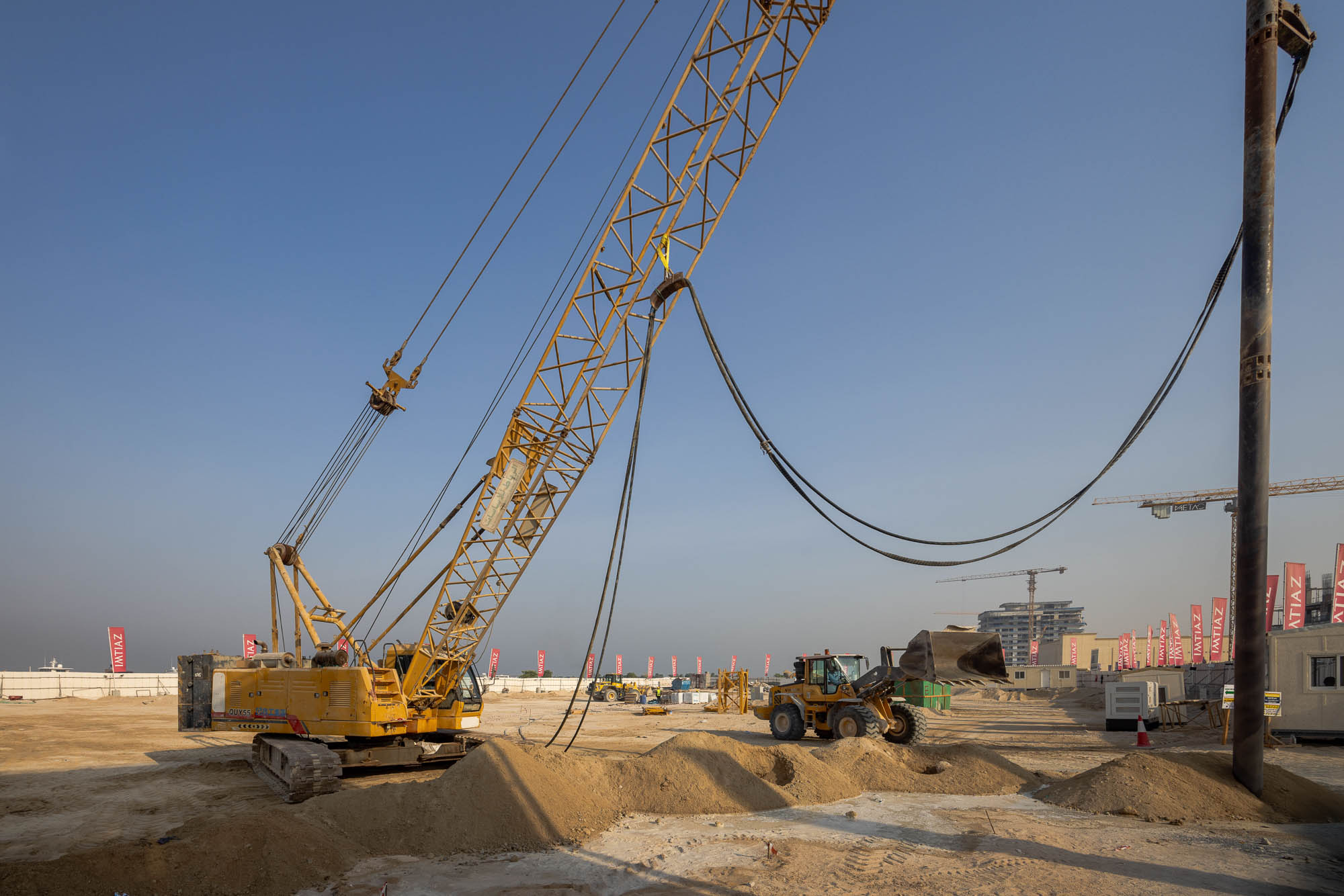 Cotier House 2 By Imtiaz A construction site featuring a yellow crane and a loader with piles of sand and cranes in the background.