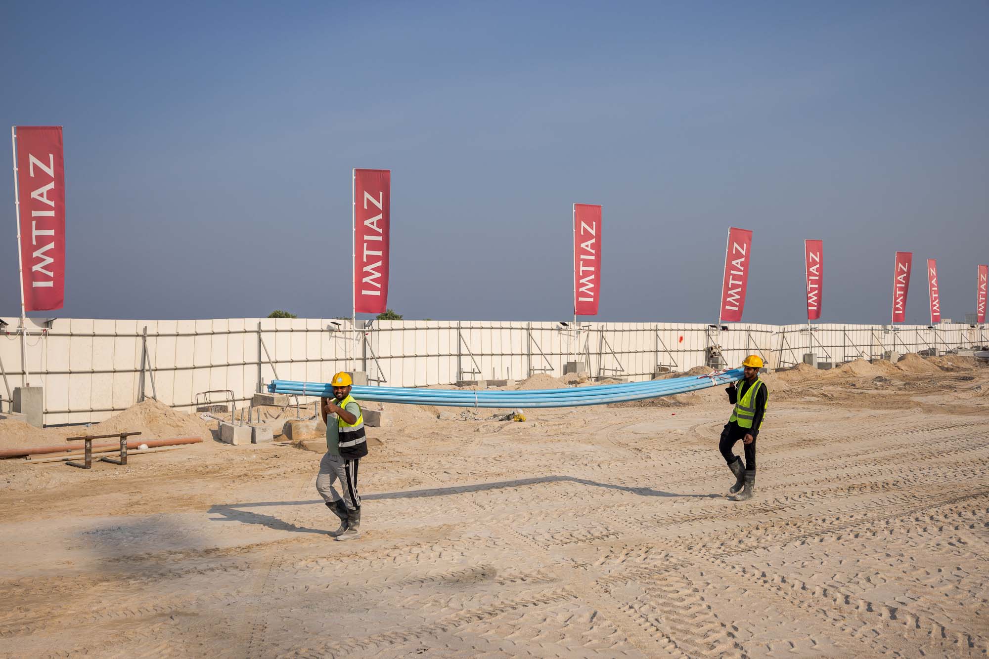Cotier House 2 By Imtiaz Two construction workers carry blue pipes on a sandy site, with banners displaying "IMTIAZ" in the background.