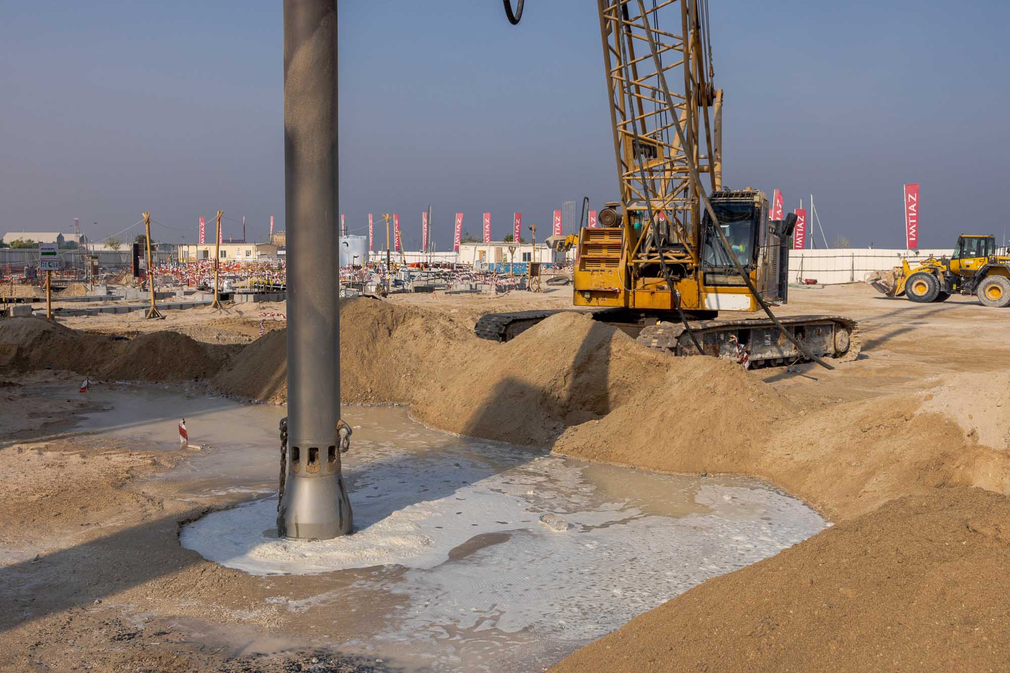 Cotier House 2 By Imtiaz Construction site with heavy machinery, a large drill, and piles of sand near a concrete foundation.