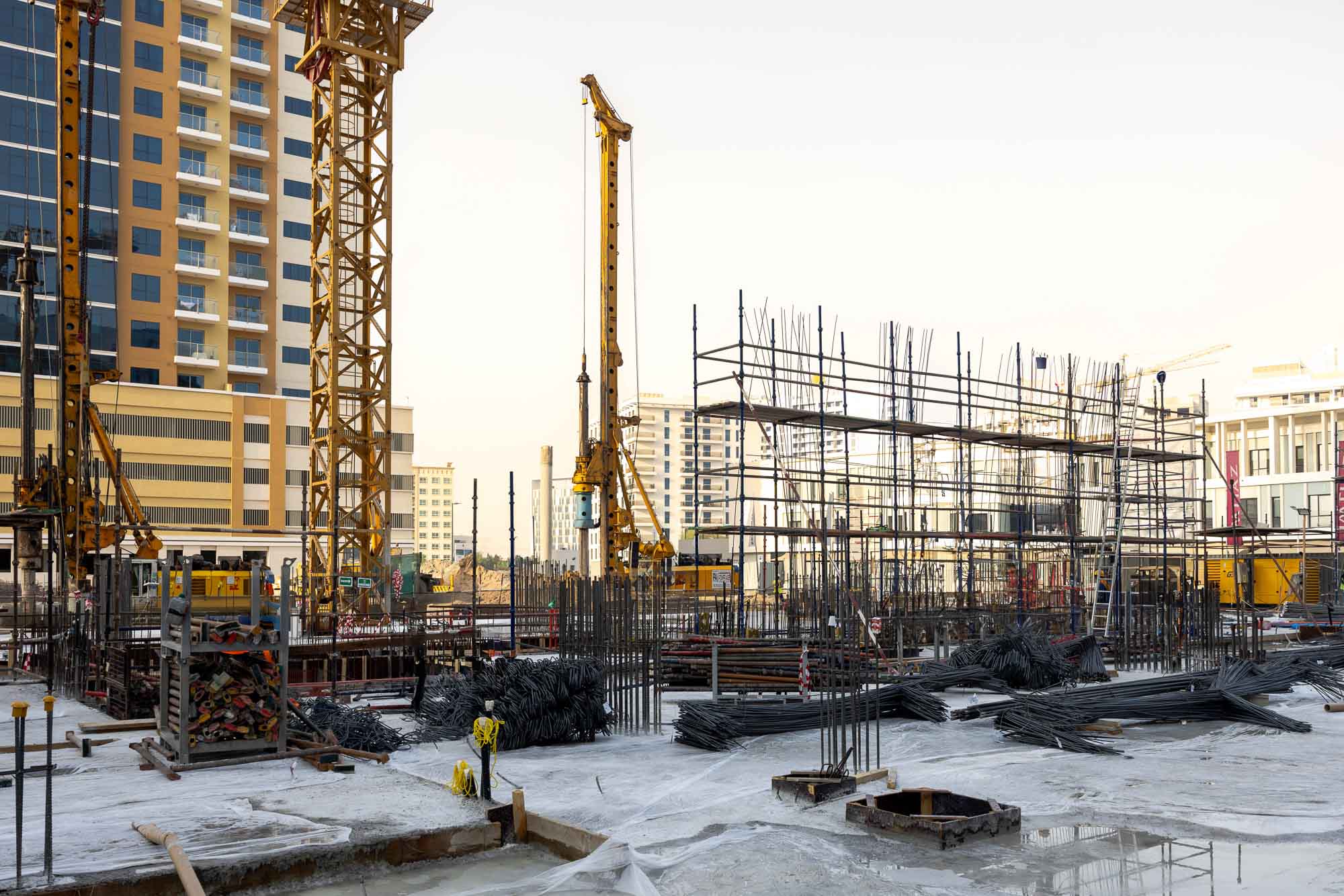 Cove Edition I By Imtiaz 1 Construction site featuring scaffolding, cranes, steel bars, and materials against a backdrop of buildings.