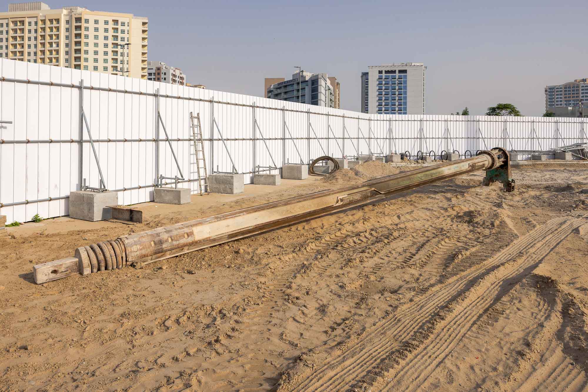 Cove Grand By Imtiaz Construction site with a large wooden structure on sandy ground, surrounded by a white fence and buildings in the background.