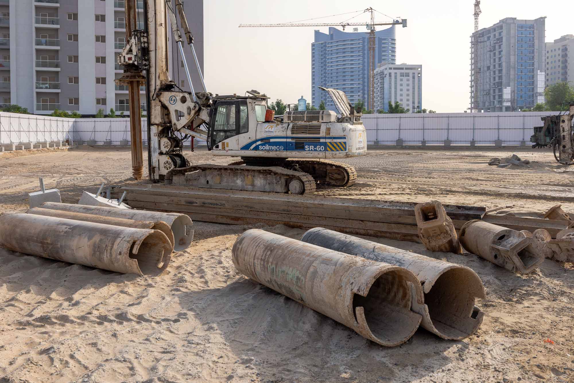 Cove Grand By Imtiaz Construction site with a drilling machine, several large cylindrical pipes, and a sandy ground. Background shows buildings.