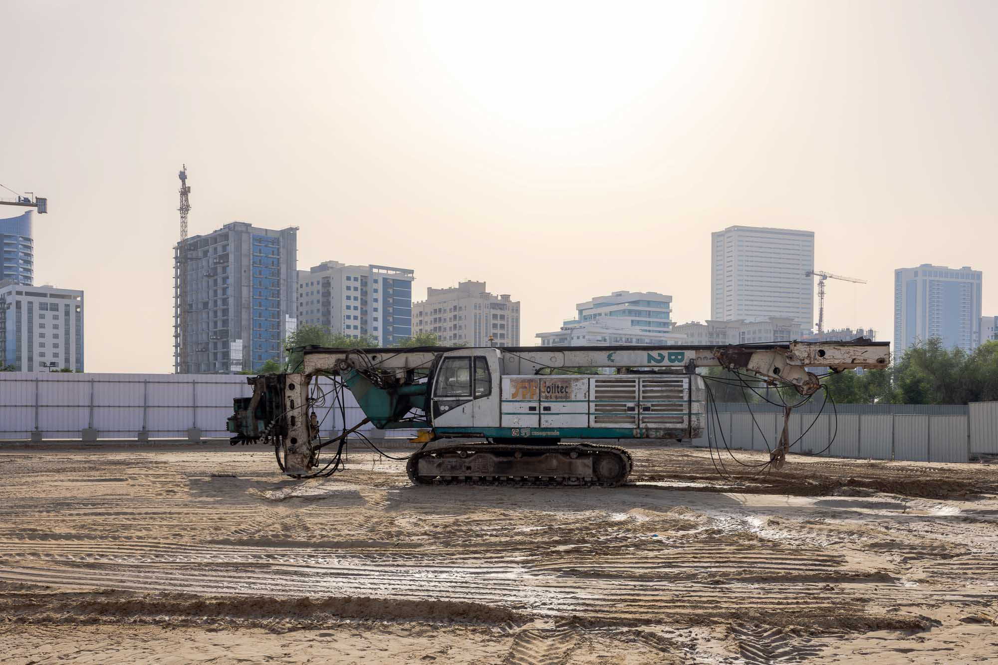 Cove Grand By Imtiaz A construction machine on a sandy lot with city buildings and cranes in the background under a clear sky.