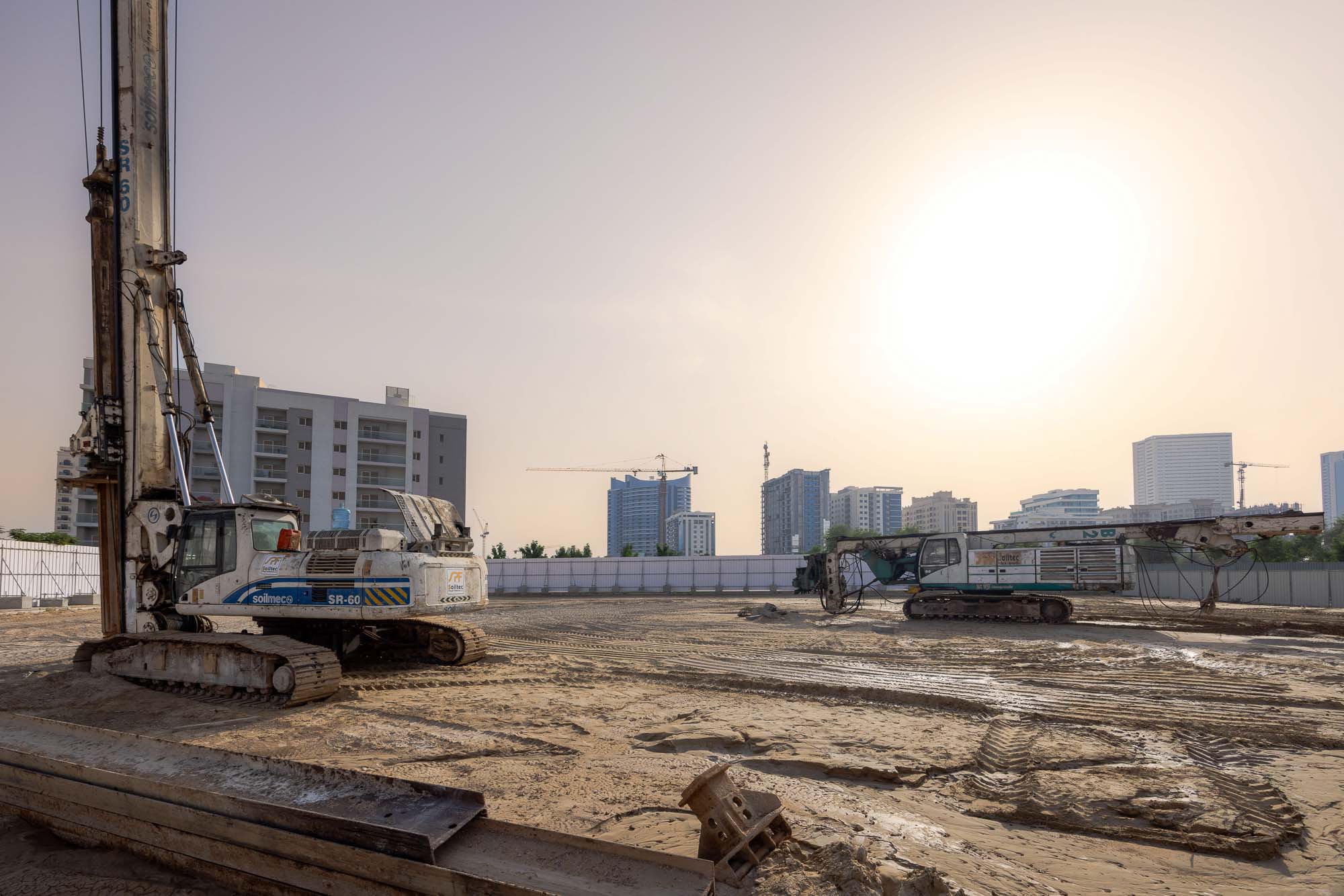 Cove Grand By Imtiaz Construction site with two large drilling machines and city buildings in the background under a hazy sky.