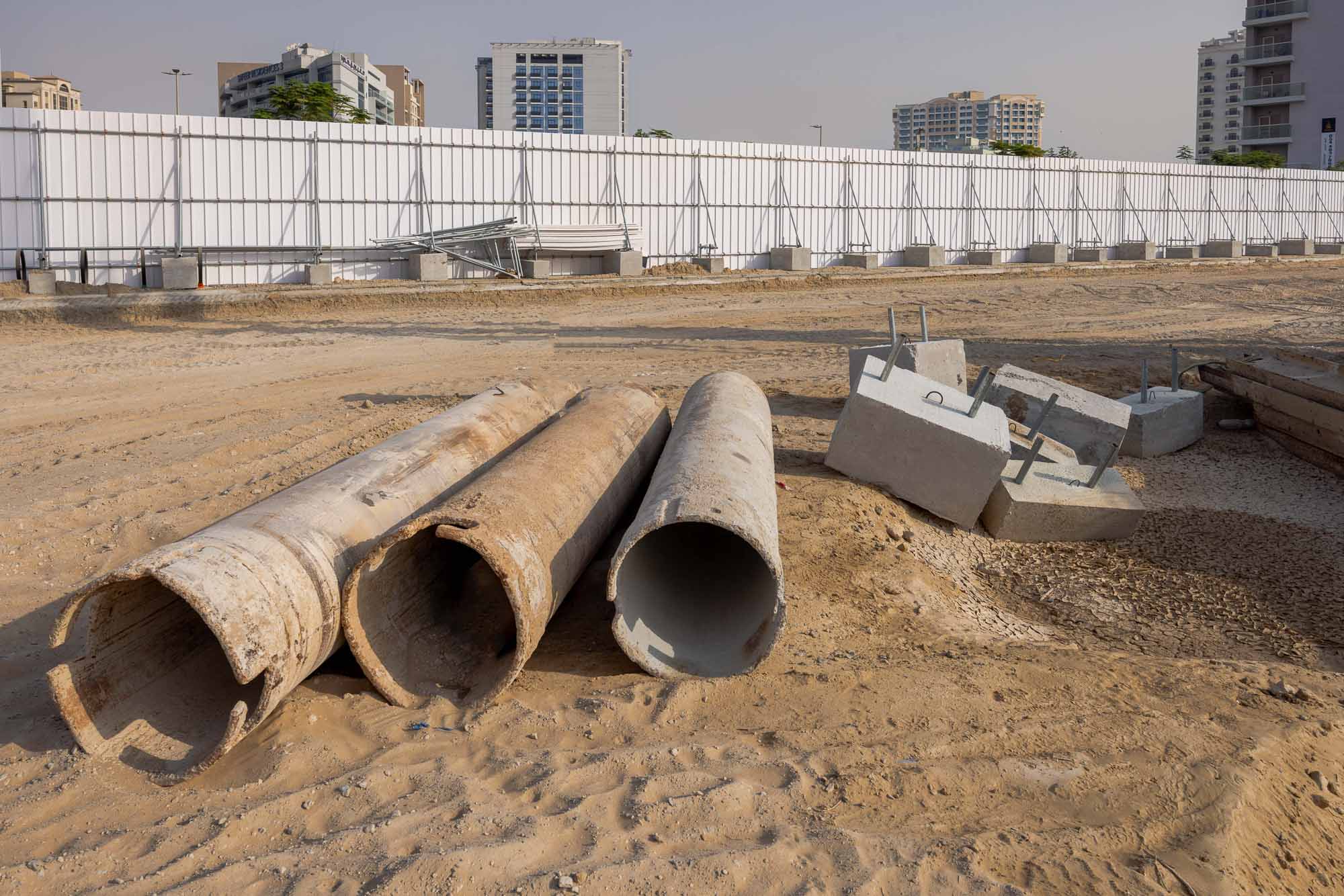 Cove Grand By Imtiaz Construction site with concrete pipes and blocks on sandy ground, surrounded by a white barricade and buildings.