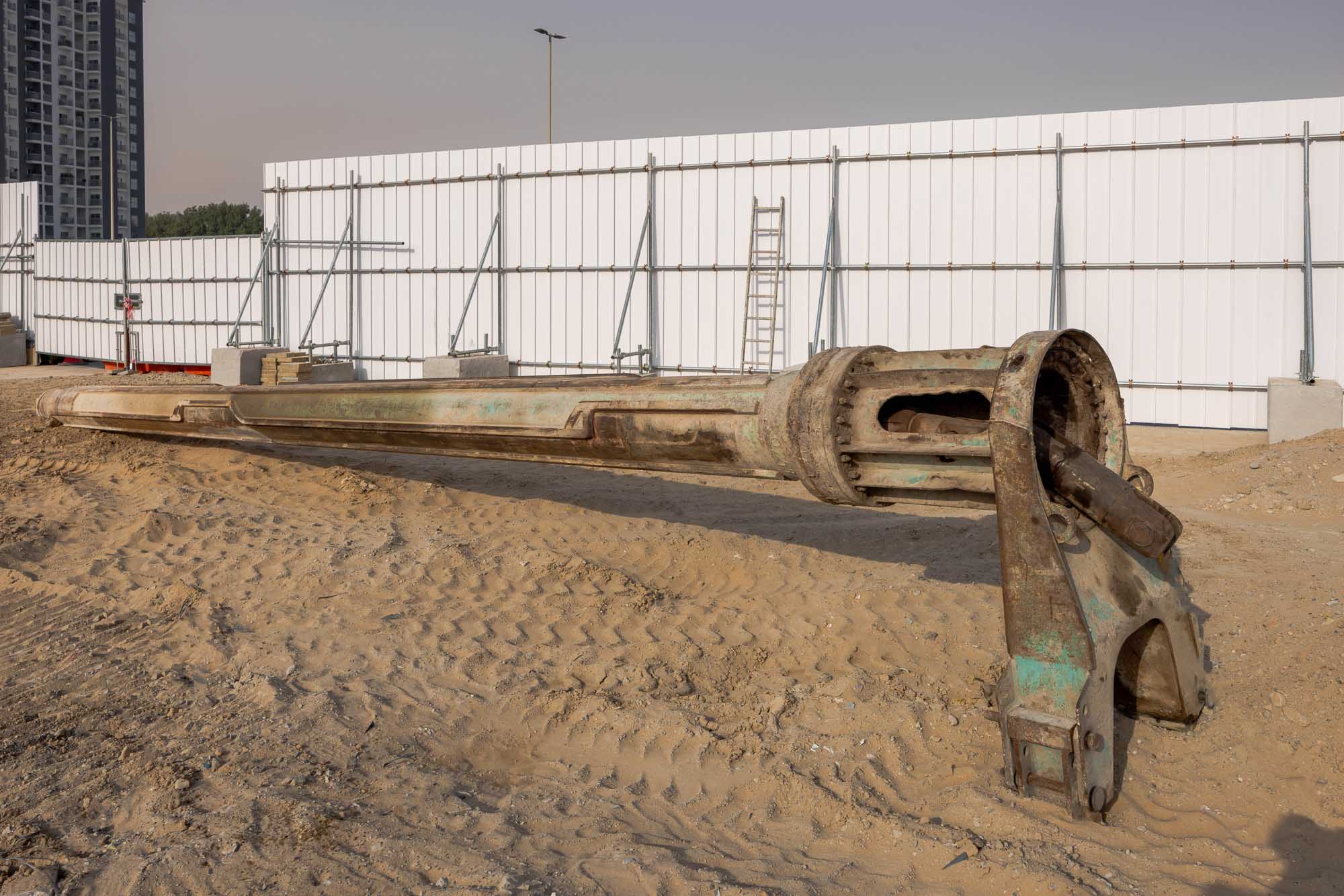 Cove Grand By Imtiaz A large, rusty construction pipe lies on sandy ground near white fencing and a metal ladder in the background.