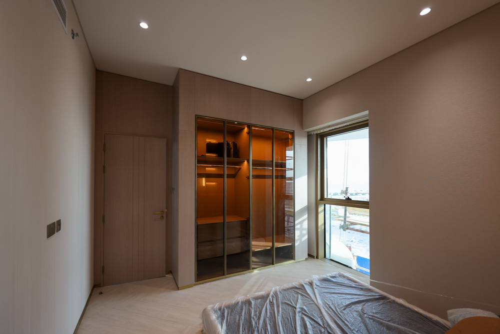 Beach Walk Residence A modern bedroom featuring a wardrobe with glass doors, a window, and soft lighting. A bed is partially covered.