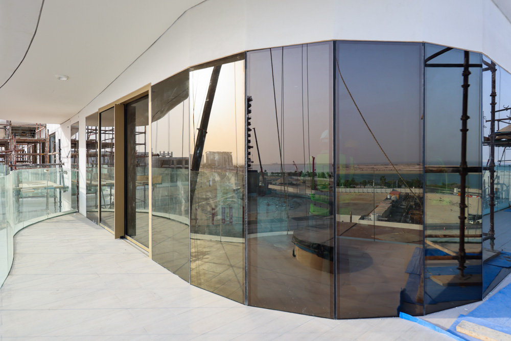 Beach Walk Residence A modern balcony with curved glass walls, overlooking a construction site and distant coastline at sunset.