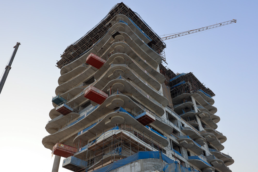 Beach Walk Iii By Imtiaz A tall building under construction, showcasing curved concrete balconies and scaffolding, against a clear sky.