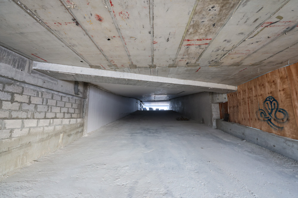 Beach Walk Iii By Imtiaz A concrete tunnel with a smooth surface, illuminated at the far end, and featuring graffiti on one wall.