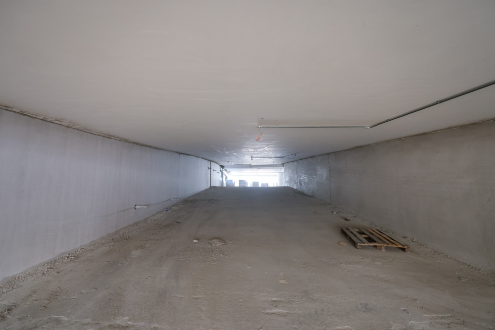 Beach Walk Iii By Imtiaz A wide, empty underground space with smooth walls, bare concrete floor, and a distant exit visible at the end.