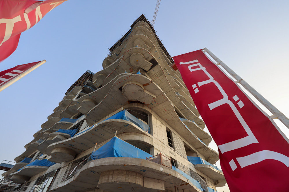 Beach Walk Iii By Imtiaz A modern building under construction with concrete layers, covered balconies, and large red banners in the foreground.